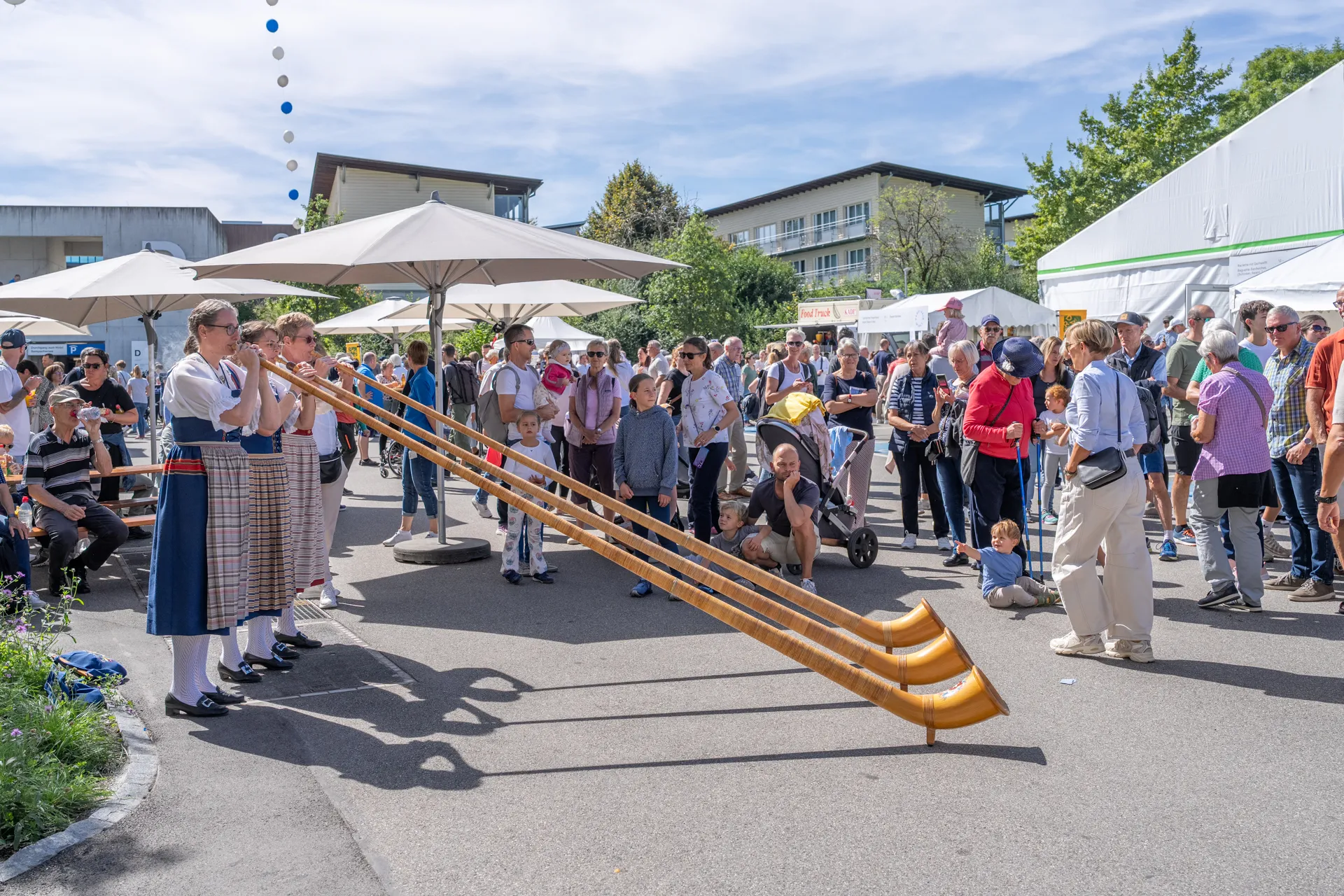 Drei Frauen spielen Alphorn an den Tagen der offenen Tür der Schweizer Paraplegiker-Gruppe. Von der Seite Fotografiert, im Hintergrund beobachten Besucherinnen und Besucher das Geschehen.