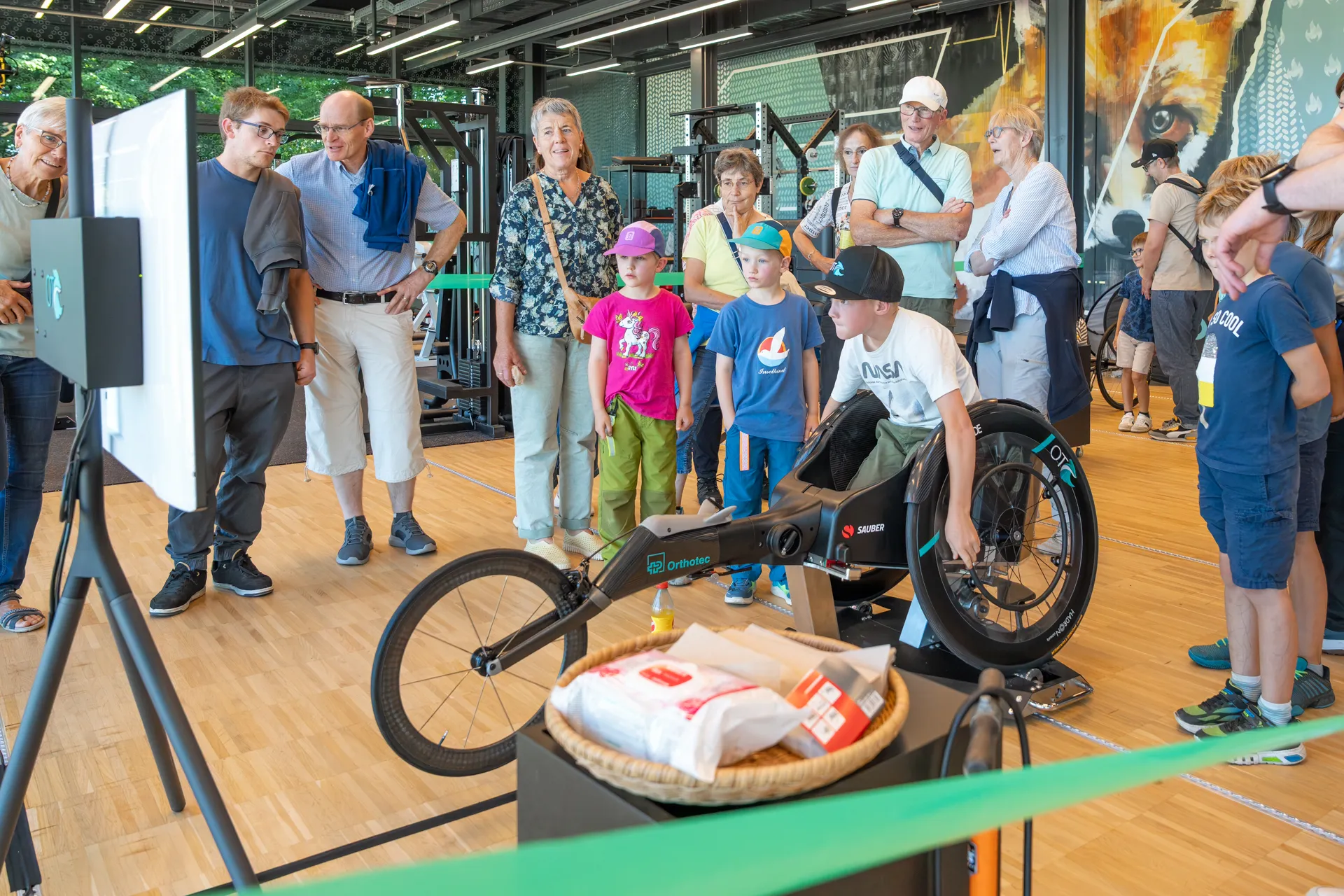 Ein Junge sitzt auf einem Trainingsrad in der Trainingshalle auf dem Campus Nottwil, während ihn weitere Besucherinnen und Besucher beobachten.