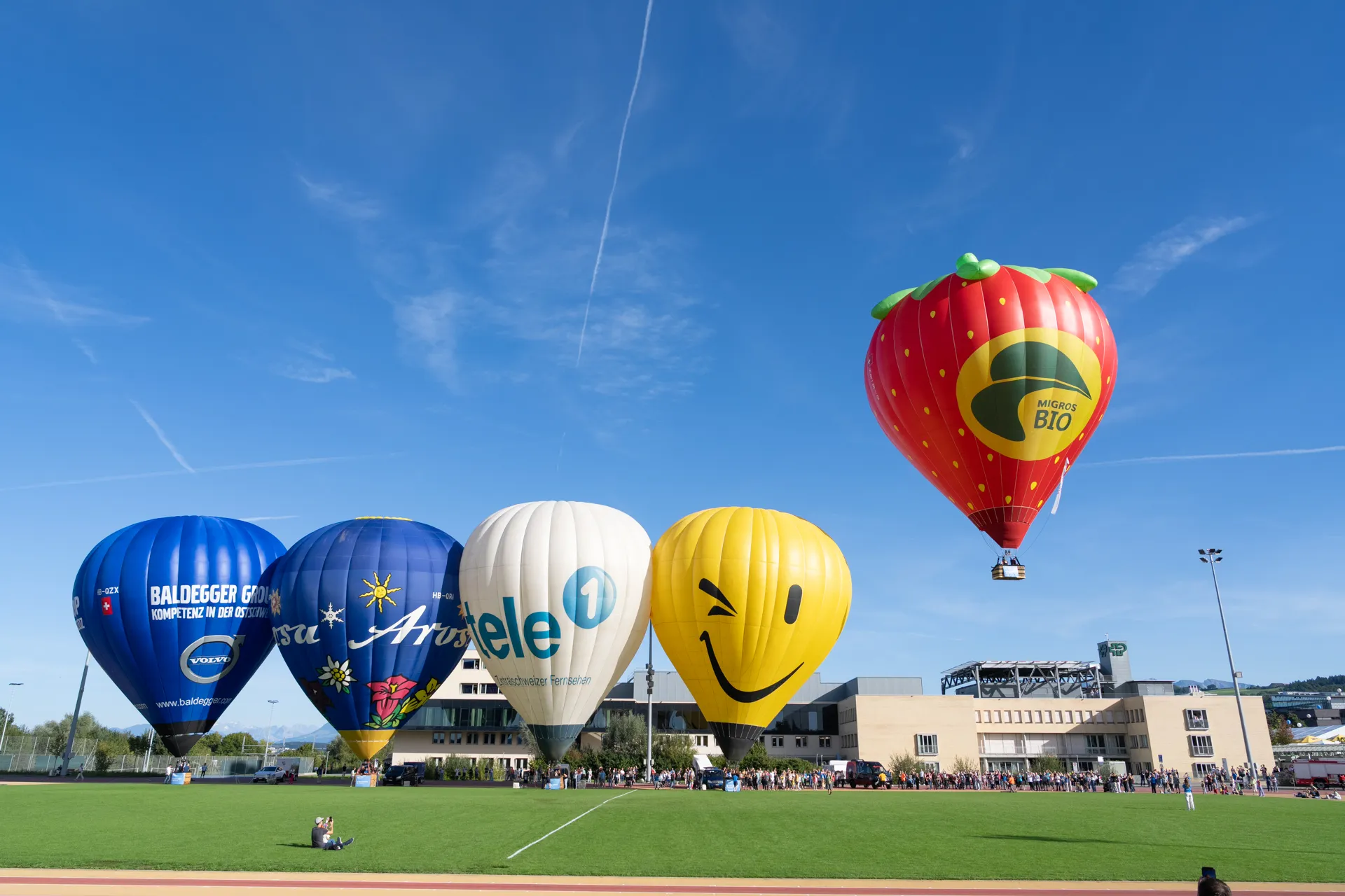 Vier Heissluftballone auf dem Sportplatz der Schweizer Paraplegiker-Gruppe vor blauem Himmel, ein Ballon hebt ab.