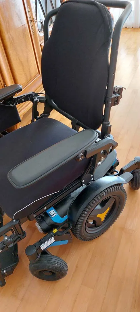 Elektrischer Rollstuhl mit blauem Rahmen und schwarzem Polster, vor einem Holzschrank stehend.