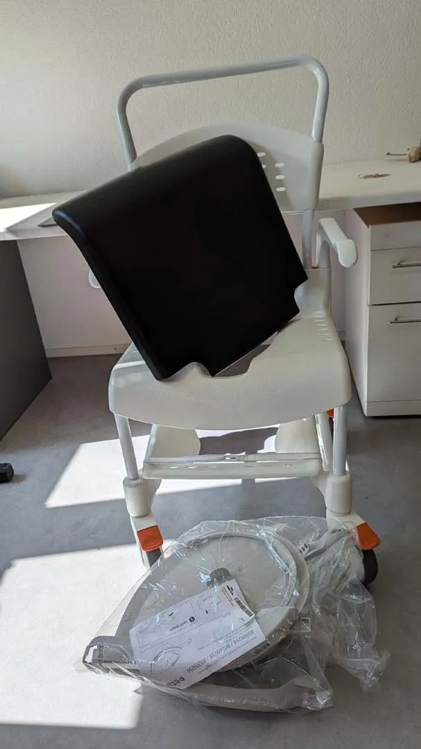 Weißer Toilettenstuhl mit schwarzem Handtuch und Waage darunter.