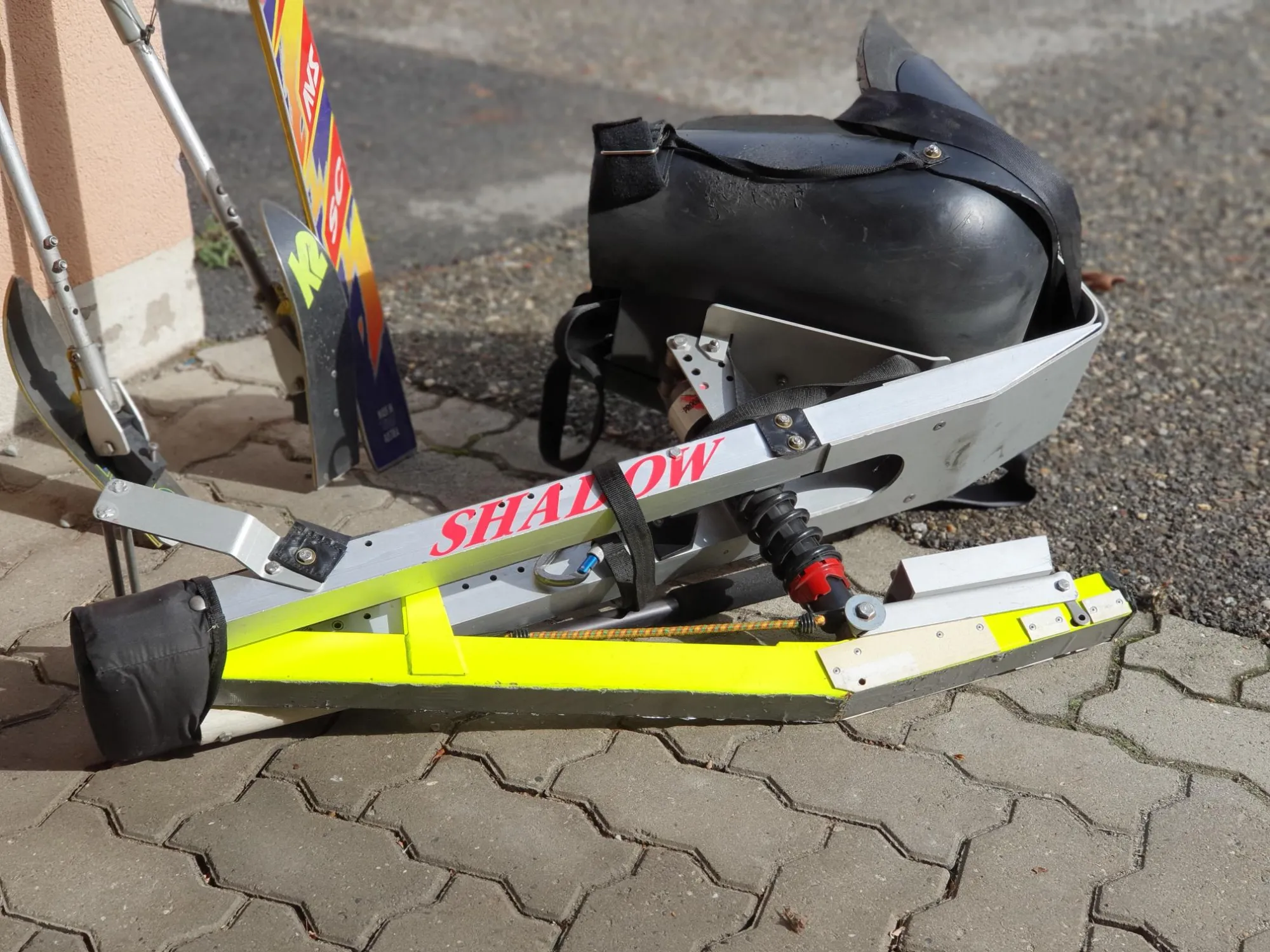 occasion-quickie-skibob-torny-01.jpg Gelbgrüne Schneeschuhe neben einem Helm und Skistock auf Pflastersteinen