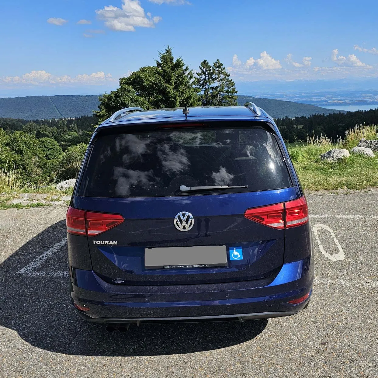 Blauer VW Touran auf Parkplatz mit Berglandschaft im Hintergrund.