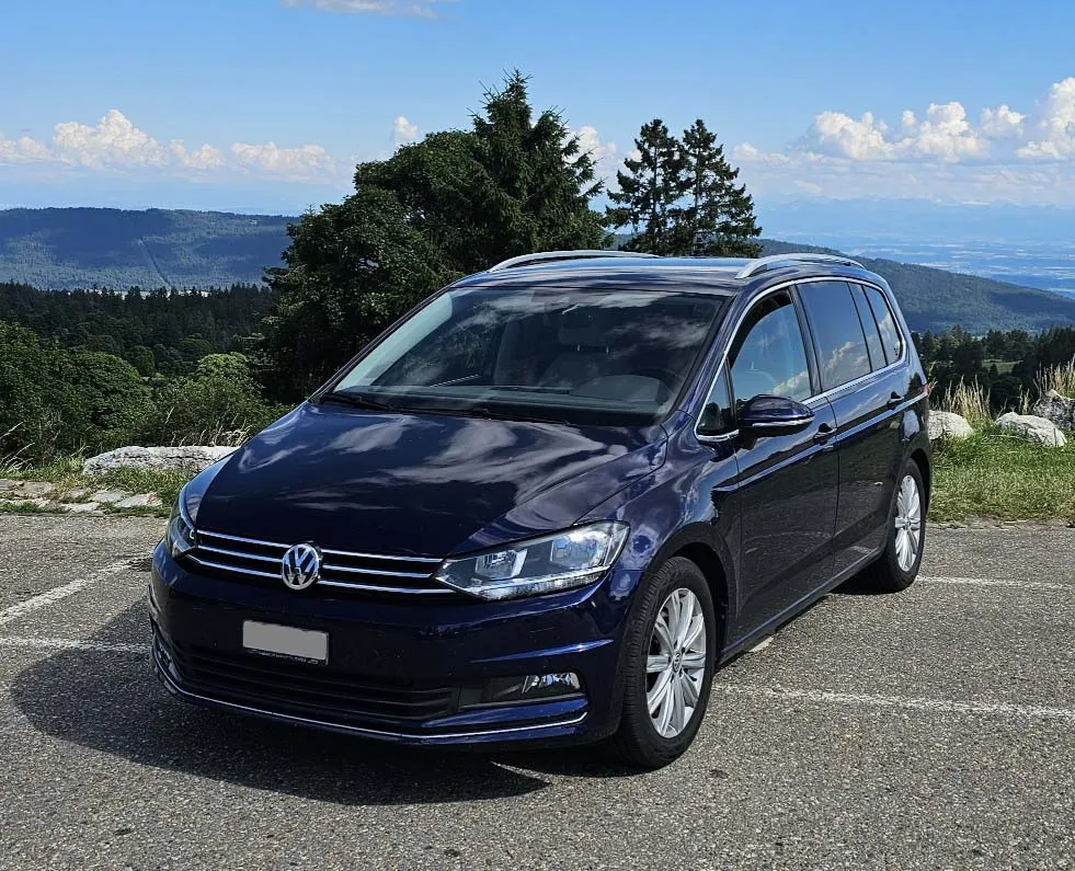 Blauer VW Sharan auf Parkplatz vor bergiger Landschaft und blauem Himmel