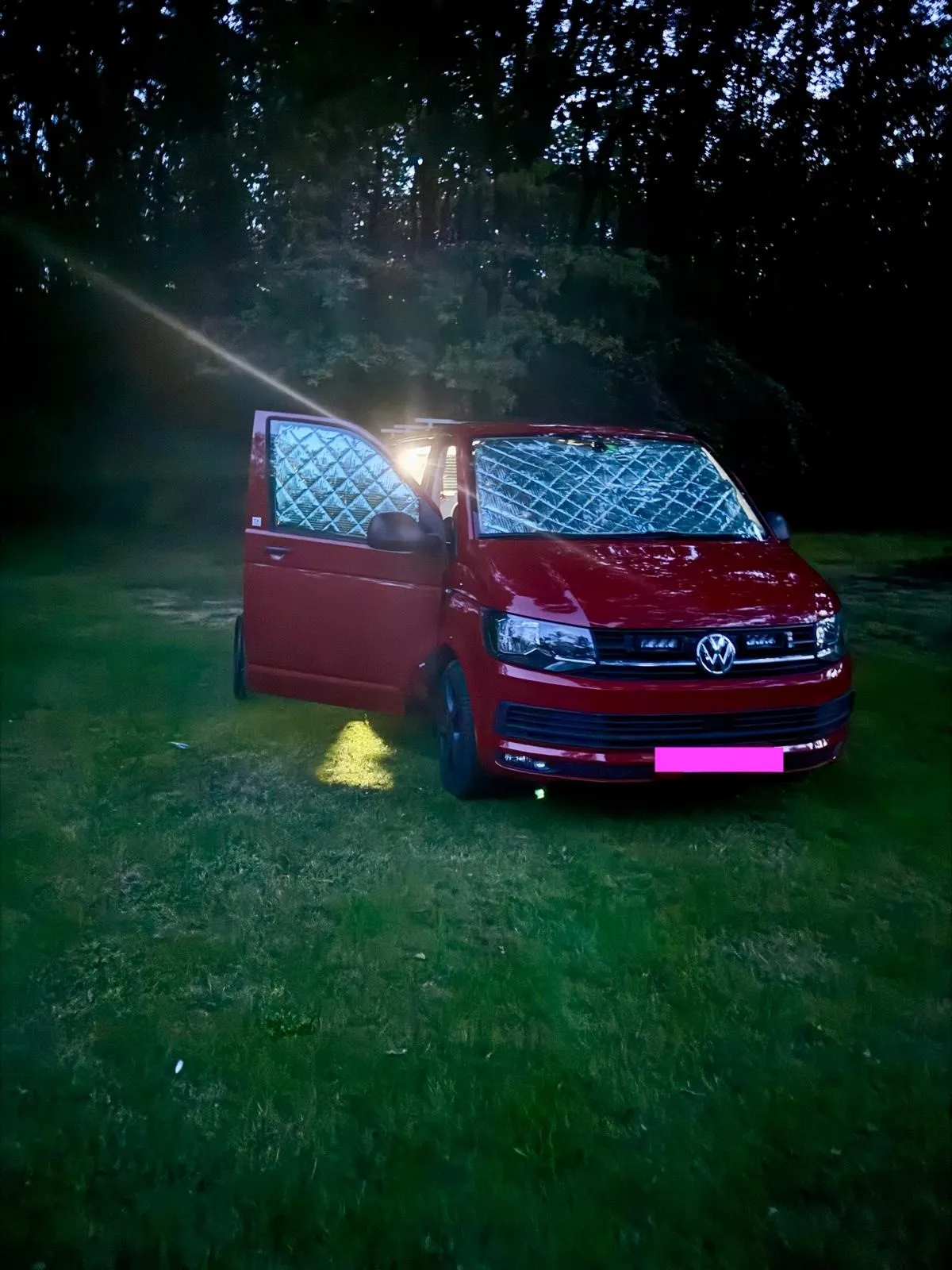 Roter Wohnwagen mit offenem Fenster im Wald bei Nacht, leuchtende Innenbeleuchtung.