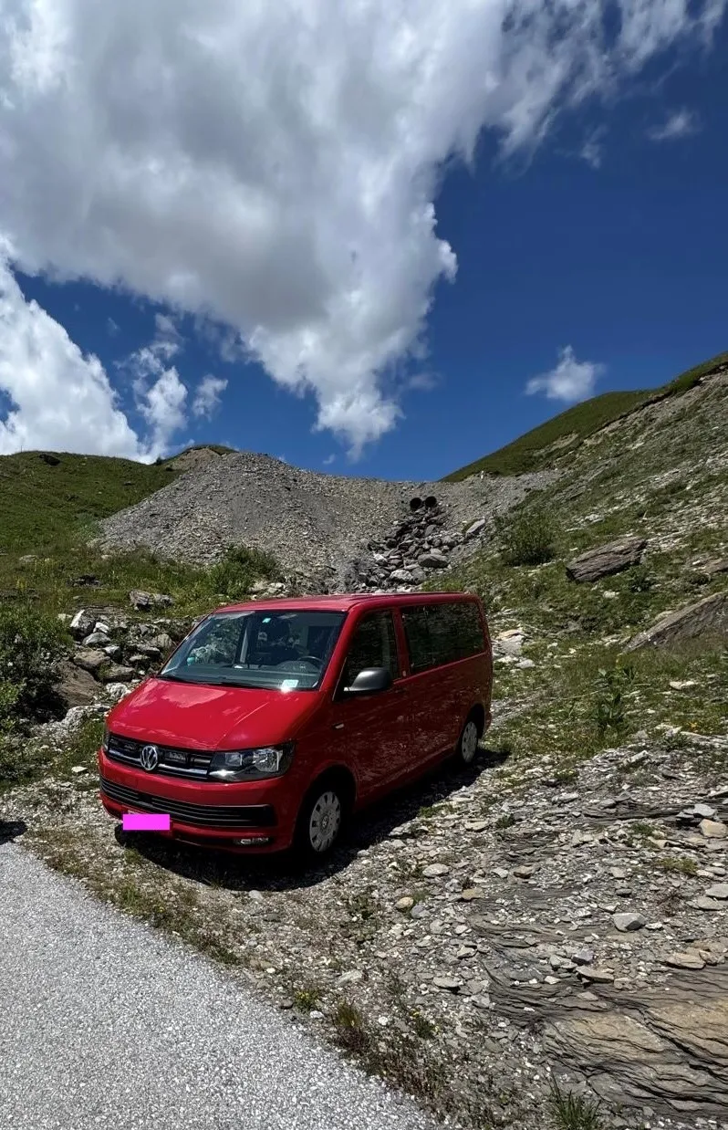 Roter Campingbus auf steiler Schotterstraße in alpiner Berglandschaft.