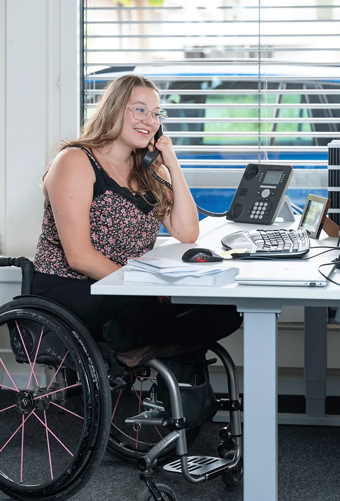 Eine Frau im Rollstuhl sitzt am Bürotisch und telefoniert.