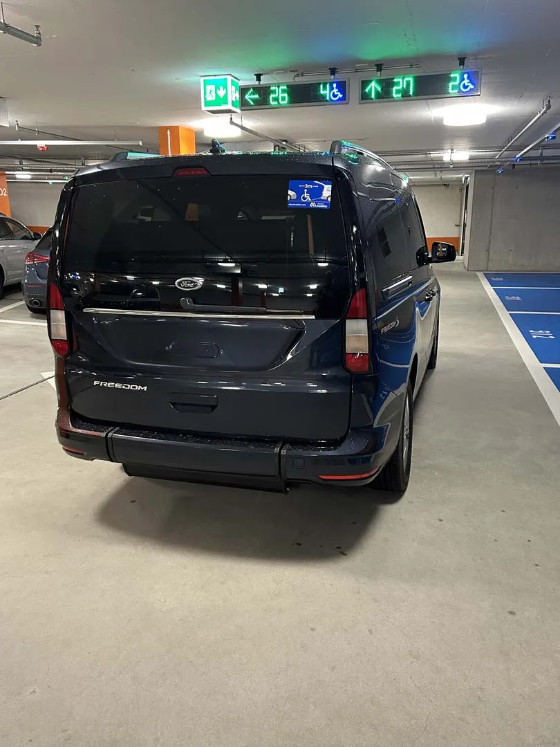 occasion-ford-tourneo-connect-blau-orthotec-04.jpg Dunkler Transporter in einem Parkhaus mit grünen Lichtsignalen an der Decke.