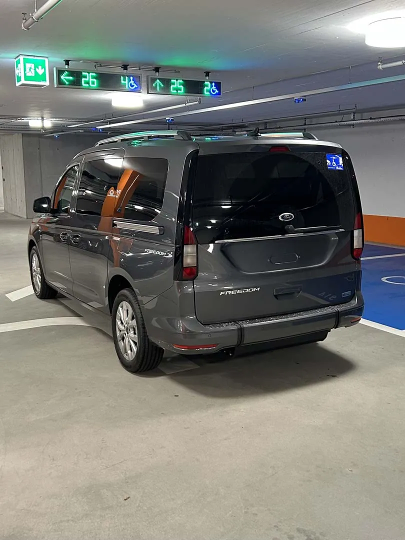 occasion-ford-tourneo-connect-grau-orthotec-02.jpg Grauer Minivan in einer Tiefgarage mit grünem Ausfahrtsschild an der Decke.
