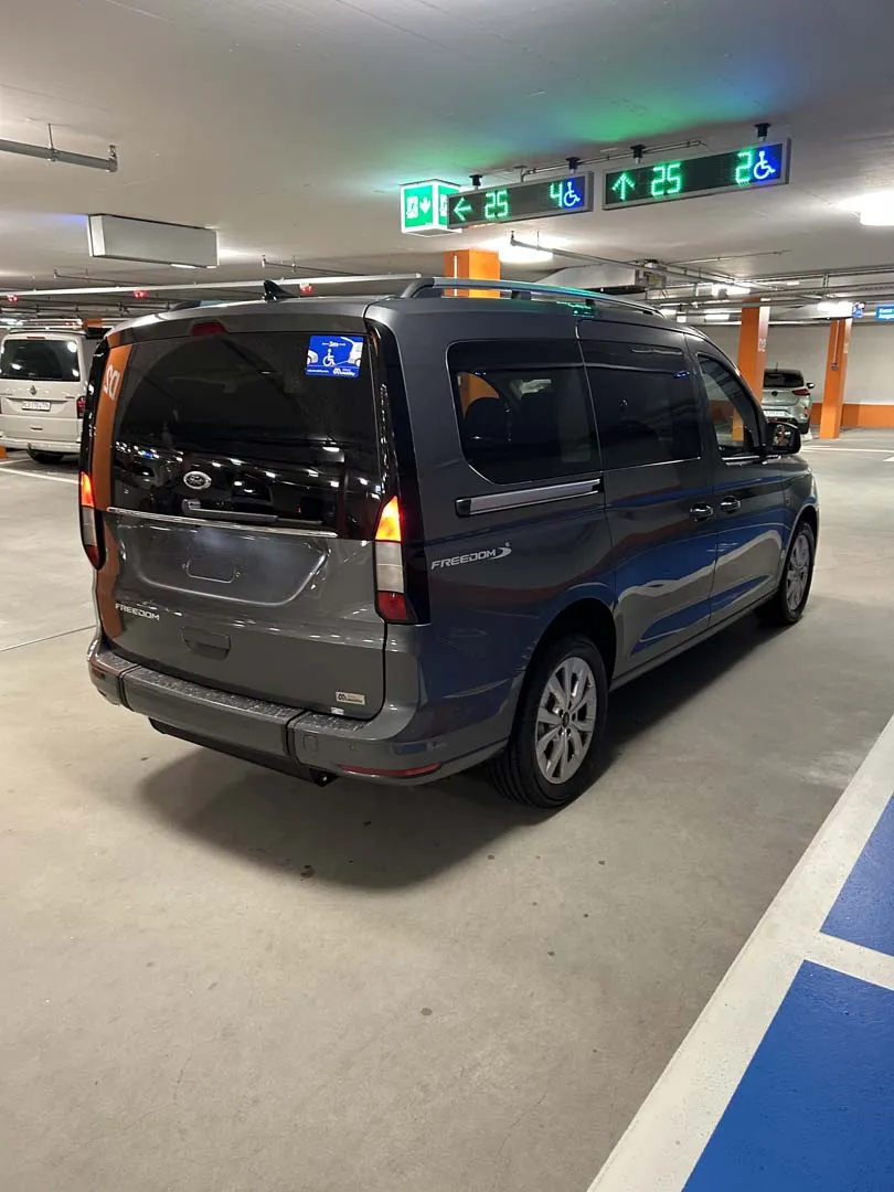occasion-ford-tourneo-connect-grau-orthotec-03.jpg Blauer Minivan in einem Parkhaus mit grünen Wegweisern an der Decke.