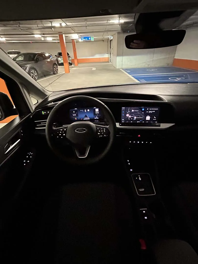 occasion-ford-tourneo-connect-grau-orthotec-05.jpg Innenansicht eines Autos mit Armaturenbrett, Lenkrad und digitaler Anzeige in einem Parkhaus.
