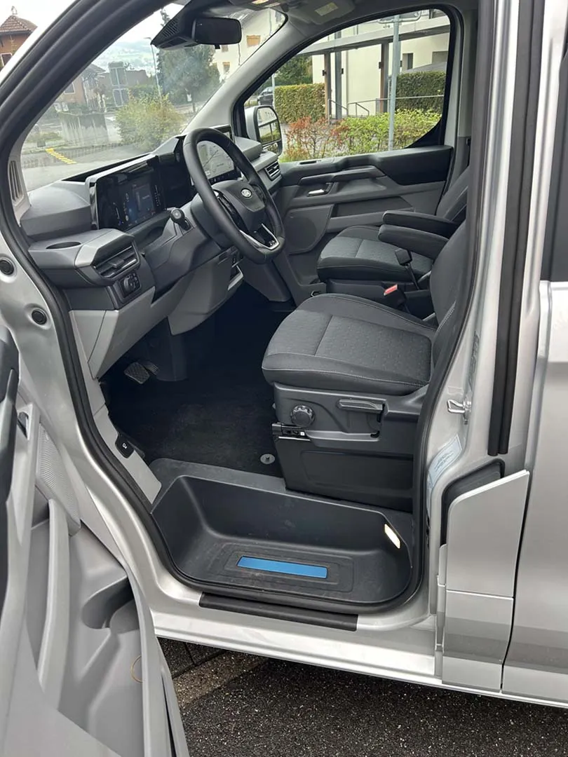 occasion-ford-tourneo-connect-silber-orthotec-03.jpg Innenansicht eines Transporter-Fahrzeugs mit geöffneter Tür, schwarzem Lenkrad und Sitzen.