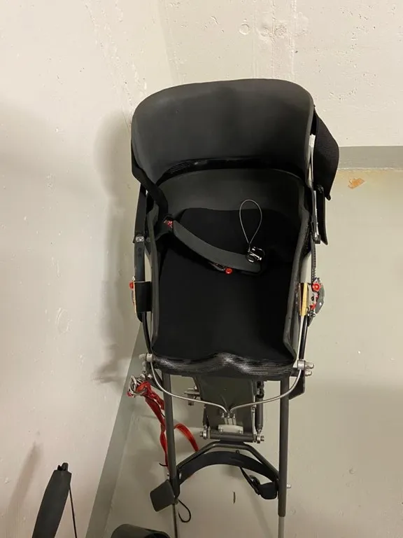 occasion-nolimit-monoskibob-sonderer-03.jpg Schwarzer Kinderwagen mit Sonnendach und rotem Gestell gegen helle Wand gelehnt.