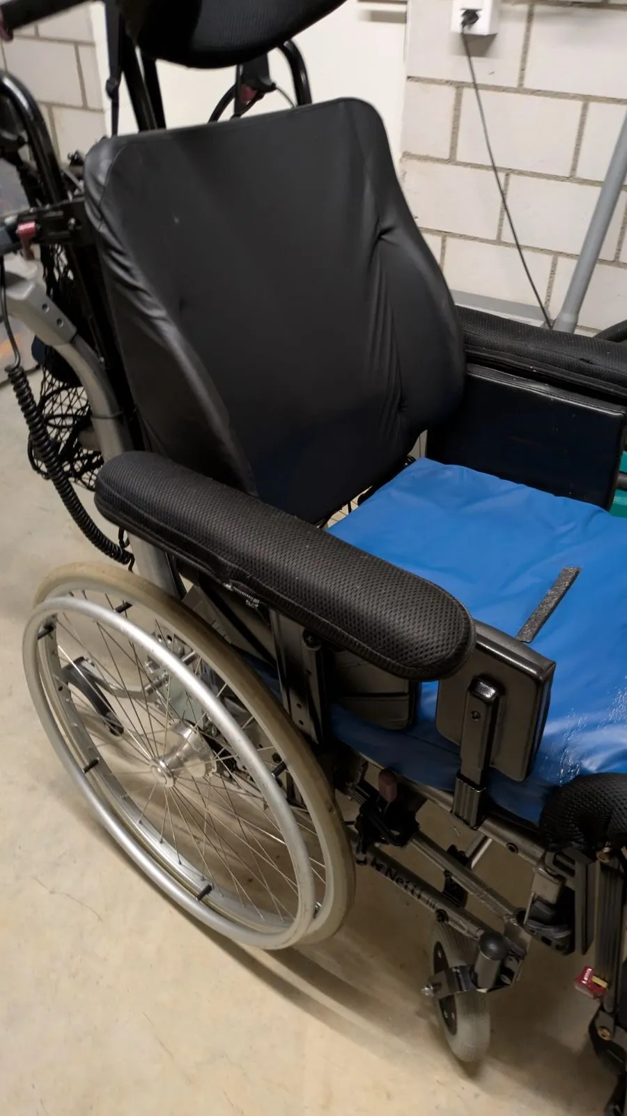 Blauer Rollstuhl mit schwarzem Sitz und Rückenlehne vor einer weißen Wand.
