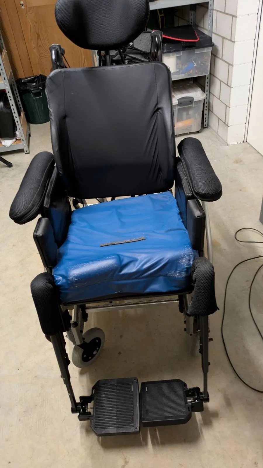 Elektrischer Rollstuhl mit schwarzer Rückenlehne und blauem Sitzkissen vor einer weißen Wand.
