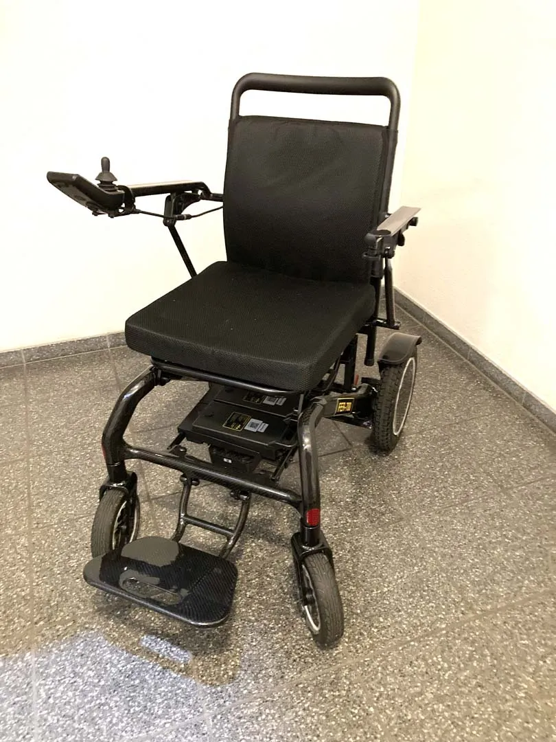 Elektrischer Rollstuhl mit schwarzem Sitz und Armlehnen auf Teppichboden.