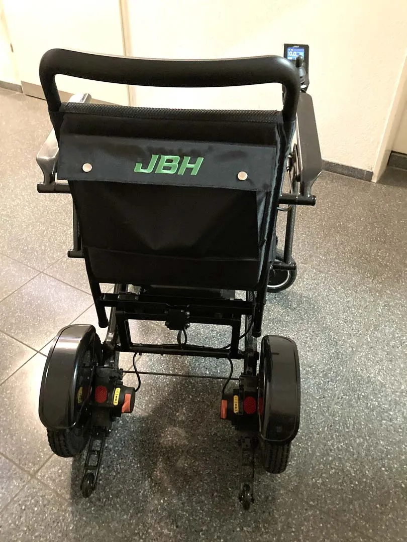 Schwarzer Rollstuhl mit grünem JBH-Logo auf der Rückenlehne, von hinten fotografiert.