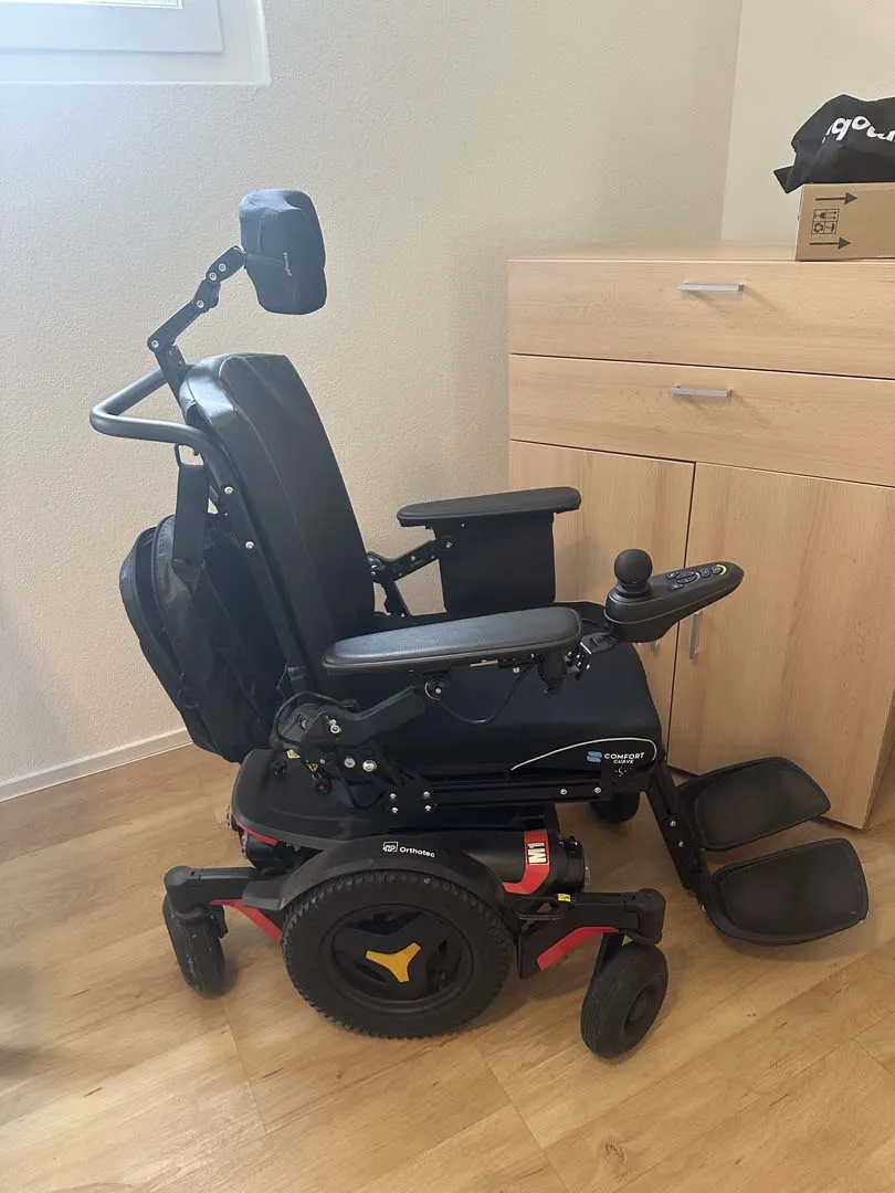 Schwarzer elektrischer Rollstuhl mit verstellbarer Kopfstütze in einem Wohnzimmer.