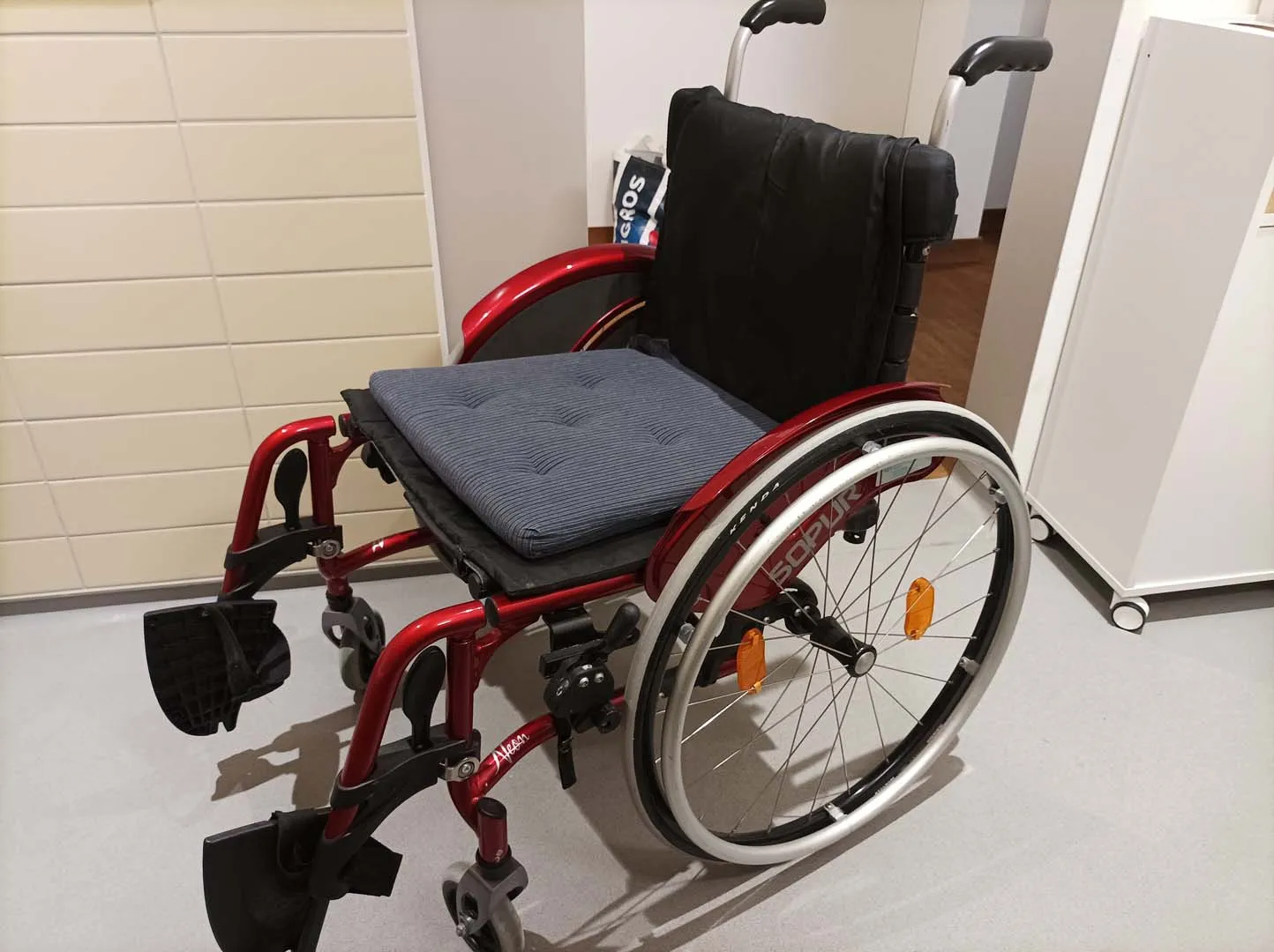 Roter Rollstuhl mit grauem Sitzpolster vor einer weißen Wand.
