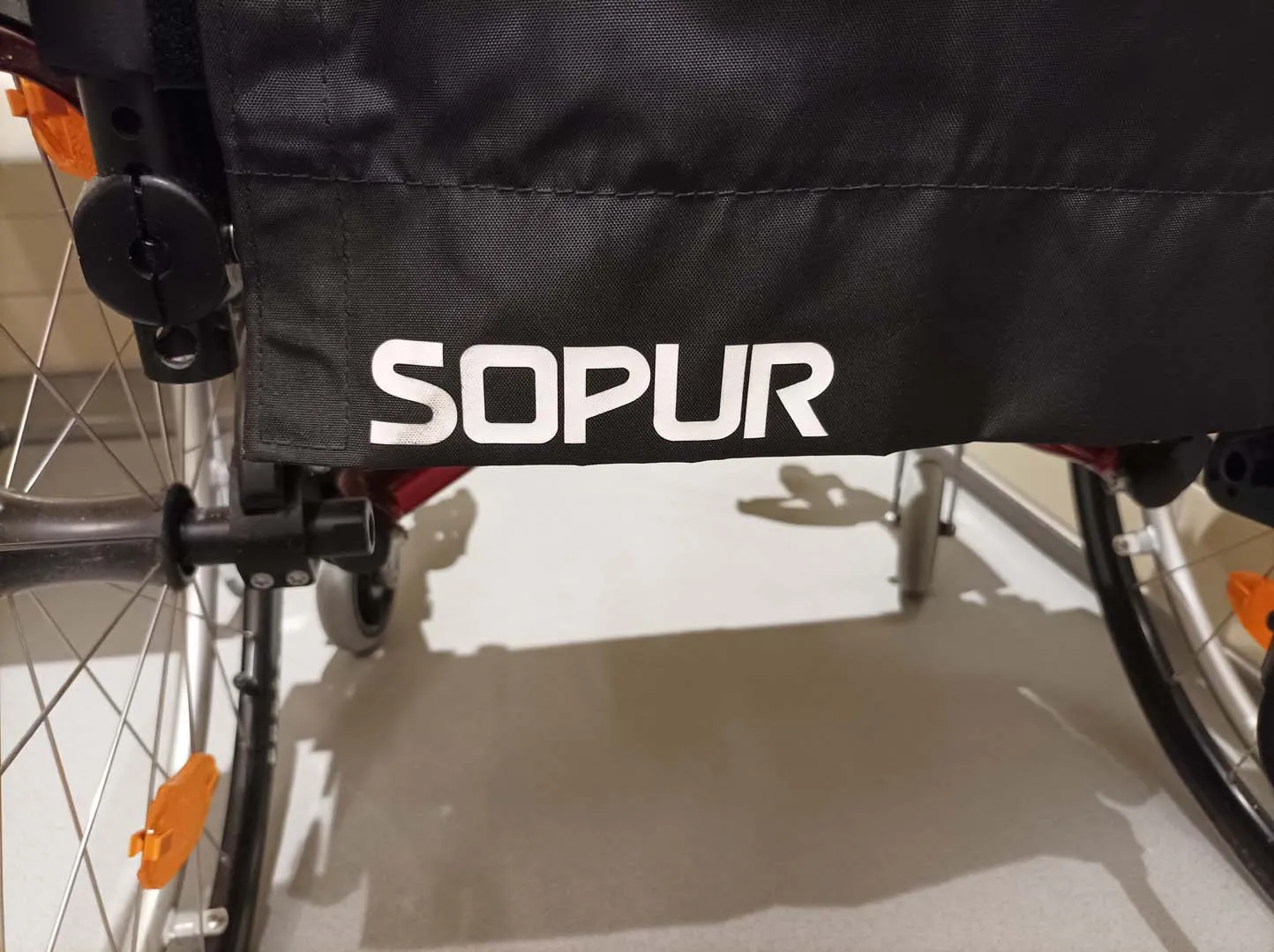 Rollstuhl mit orangefarbenen Details und dem Namen "SOPUR" auf der Rückenlehne.
