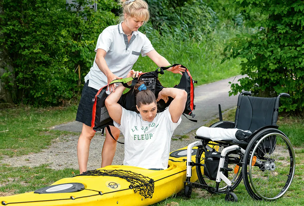 Alexia Giudi sitzt an Land in einem Kajak, neben ihr steht ihr Rollstuhl. Sporttherapeutin Amélie Fetzer unterstützt sie beim Anziehen der Schwimmweste.