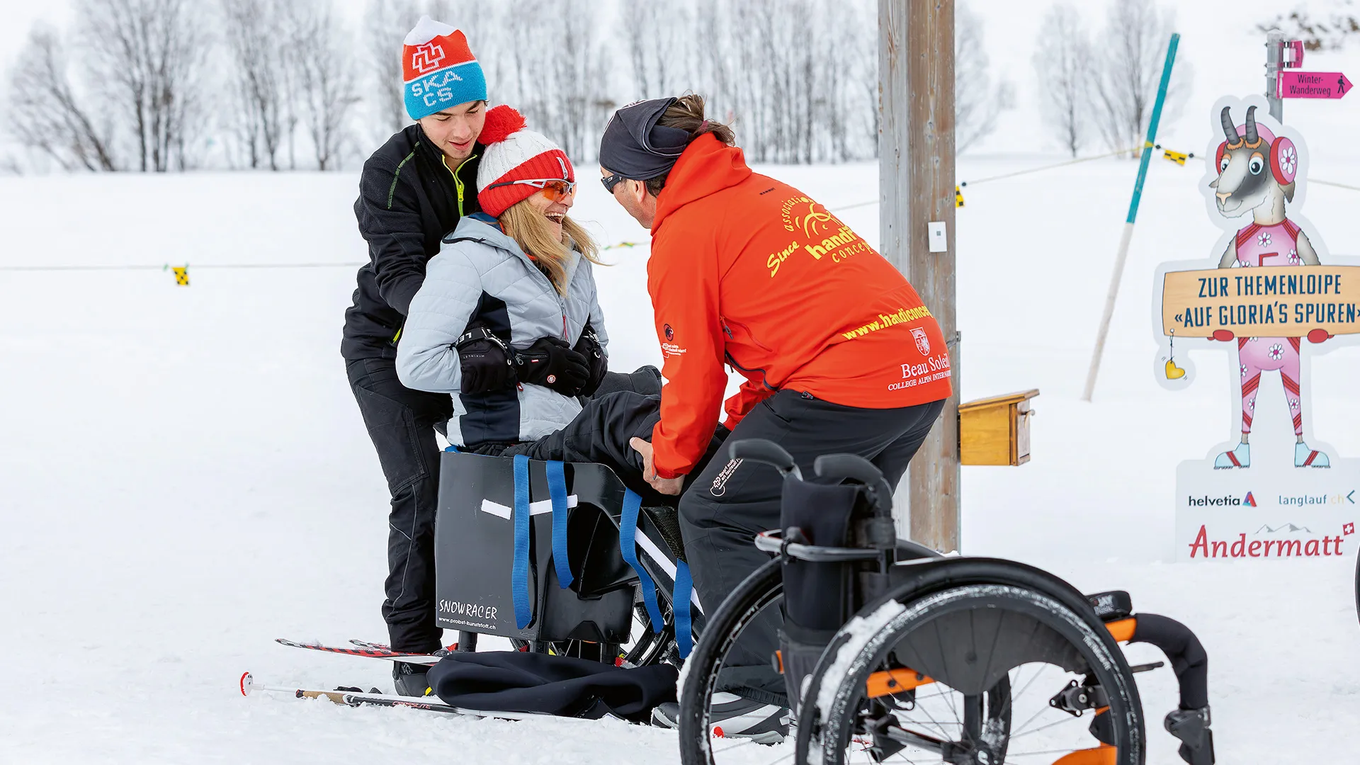 Zwei Personen unterstützen Gabriela beim Transfer vom Rollstuhl in den Langlaufschlitten.