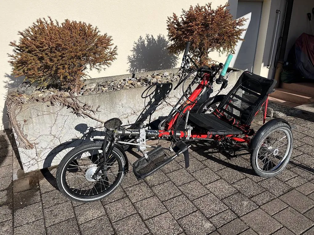 occasion-hasebike-ammann-04.jpg Rot-schwarzes Liegerad mit drei Rädern vor einer Hauswand, neben zwei Zierbäumen.