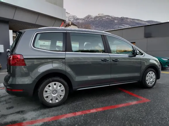 Grauer Minivan der Marke Volkswagen, geparkt auf einem Parkplatz vor Gebäuden und Bergen.