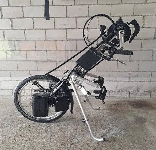 occasion-stricker-elektro-vorspannbike-kaelin-01.jpg Umgestürzter Rollstuhl mit angebautem Elektroantrieb vor einer weißen Ziegelwand.