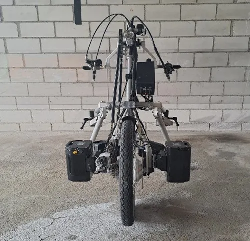 occasion-stricker-elektro-vorspannbike-kaelin-03.jpg Schwarzes Dreirad-Fahrrad mit Stützrädern und Gepäckträgern vor einer Ziegelwand.