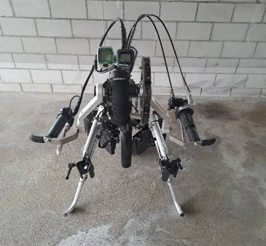 occasion-stricker-elektro-vorspannbike-kaelin-04.jpg Vierbeiniger Roboter mit Kabeln und Sensoren vor einer weißen Mauer.