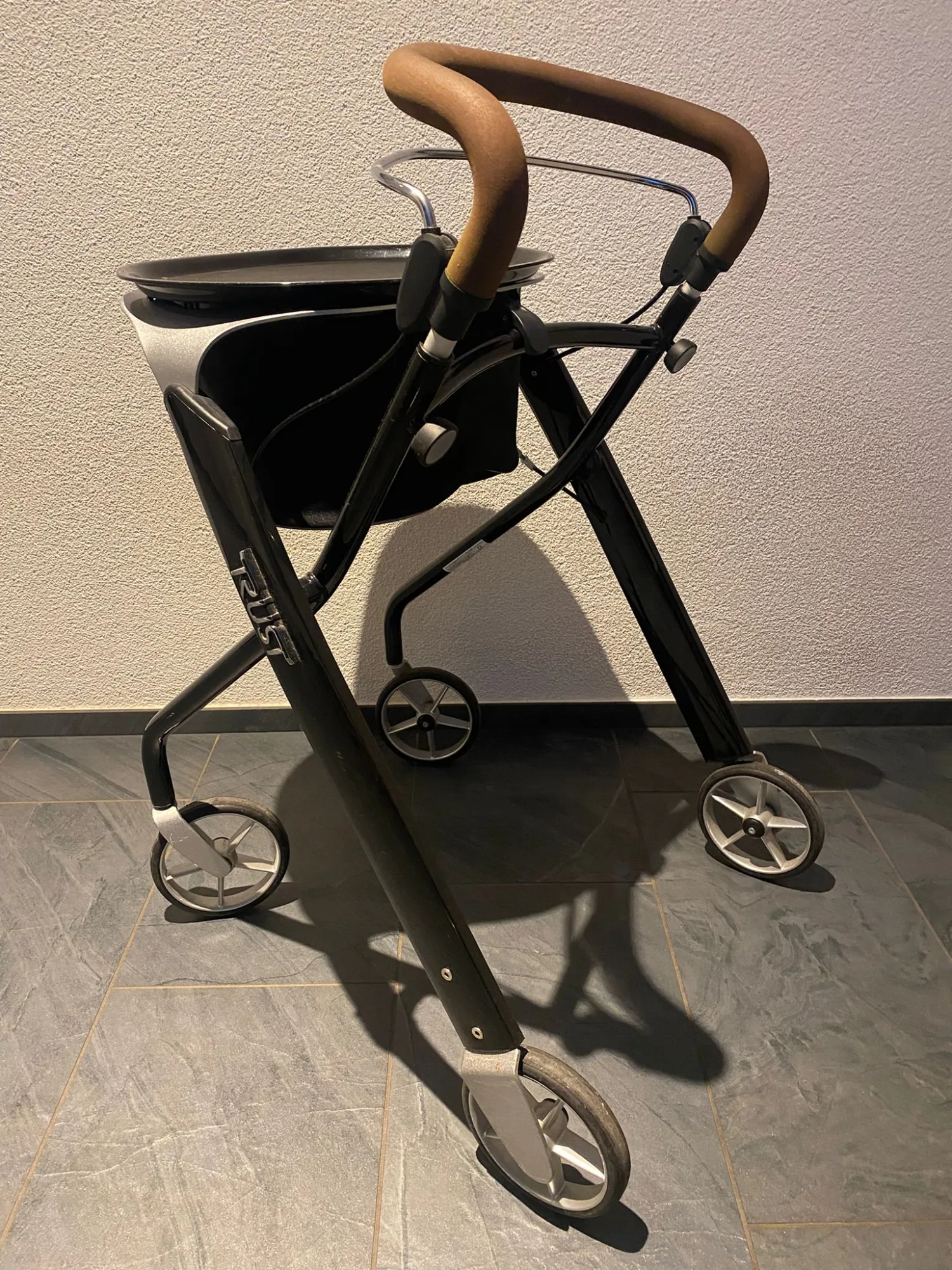 occasion-rollator-trust-weiss-01.jpg Schwarzer Rollator mit Holzgriff und Sitzfläche auf hellem Boden.