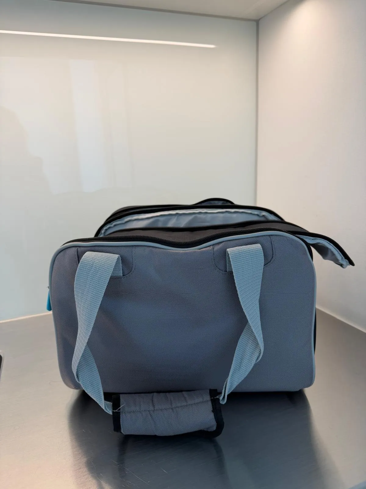 occasion-cough-assist-ofner-10.jpg Graue Sporttasche mit schwarzen Riemen auf glänzender Oberfläche vor weißer Wand.