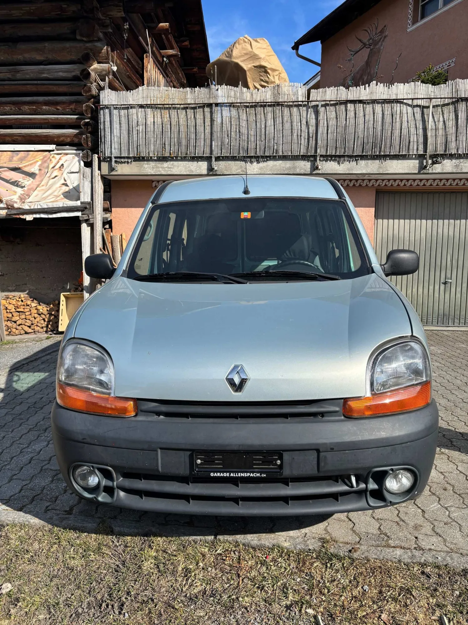 Blauer Renault Kangoo vor altem Holzschuppen und Steinhaus.