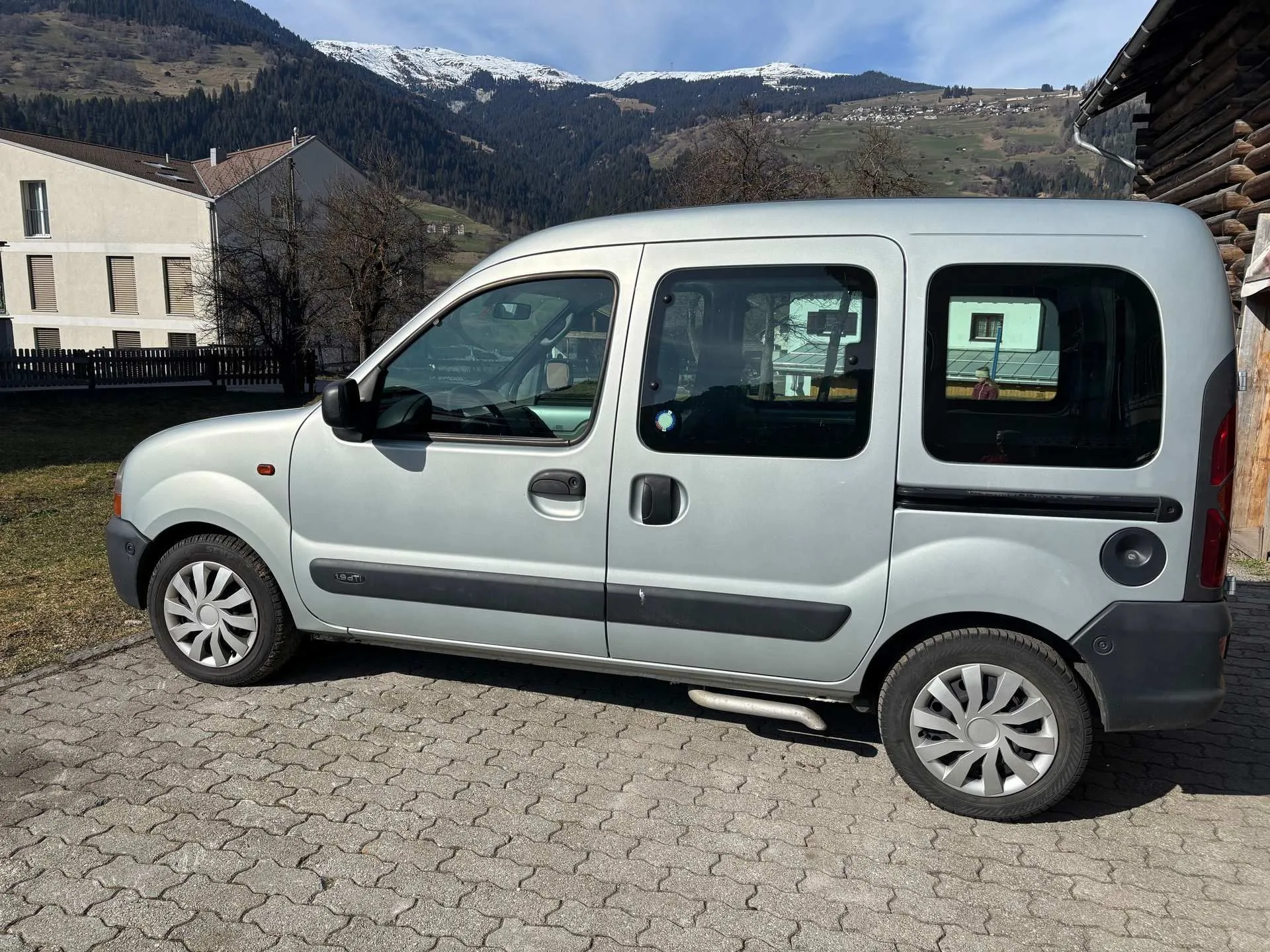 Silberner Renault Kangoo parkt auf gepflastertem Platz vor Haus, Berge im Hintergrund.