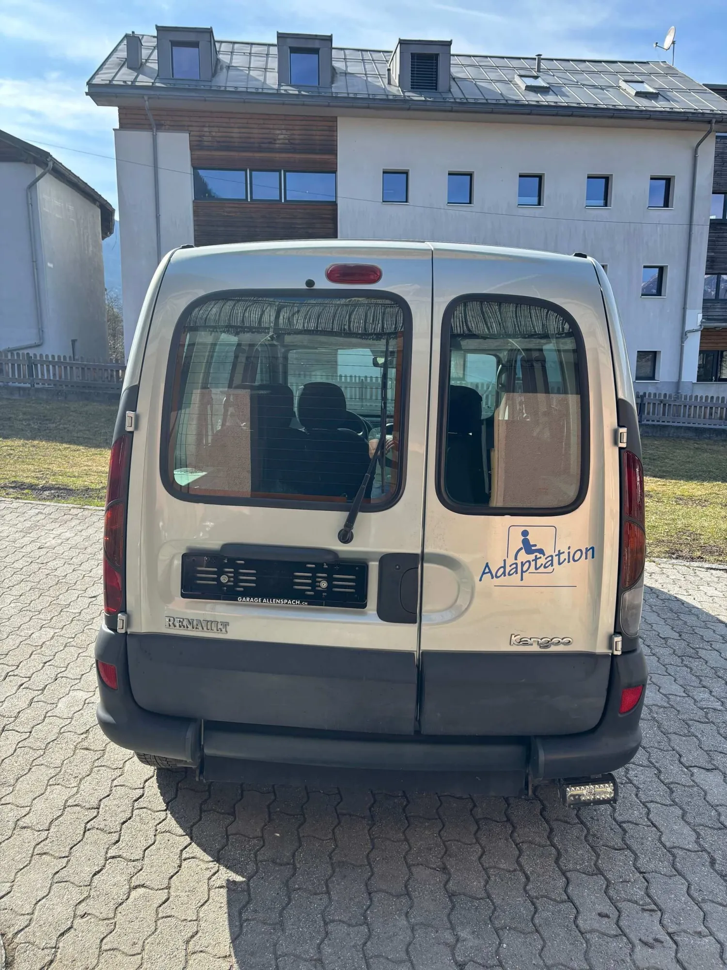 Beiger Lieferwagen mit zwei Hecktüren, geparkt vor einem weißen Gebäude.