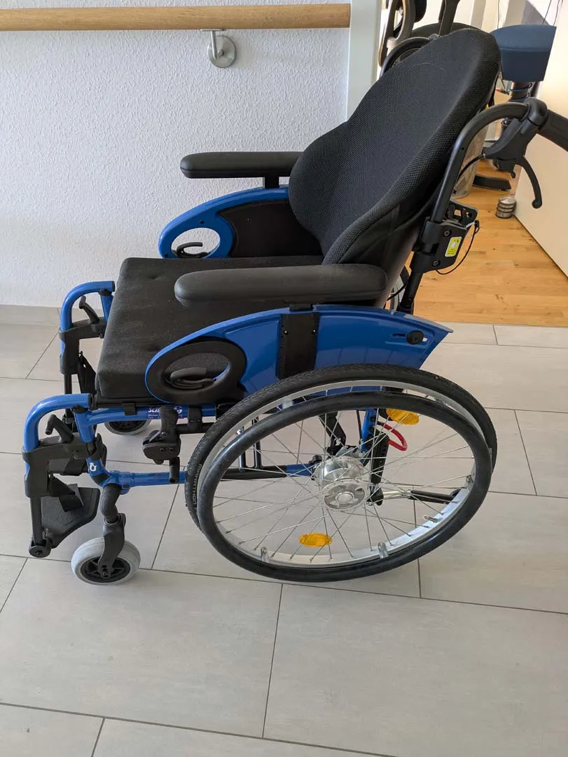 Blauer Rollstuhl mit verstellbarer Rückenlehne und Fußstützen auf Holzboden.