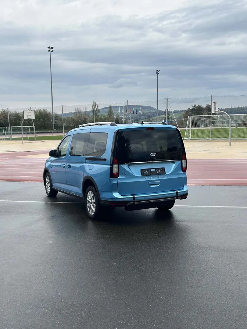 Blauer Minivan auf leerem Parkplatz vor Sportplatz bei bewölktem Himmel.