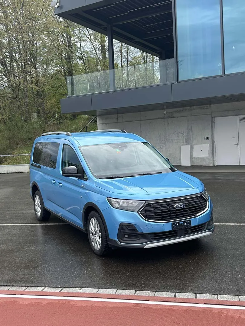 Blauer Ford Tourneo Connect Minivan auf einem Parkplatz vor einem modernen Gebäude.