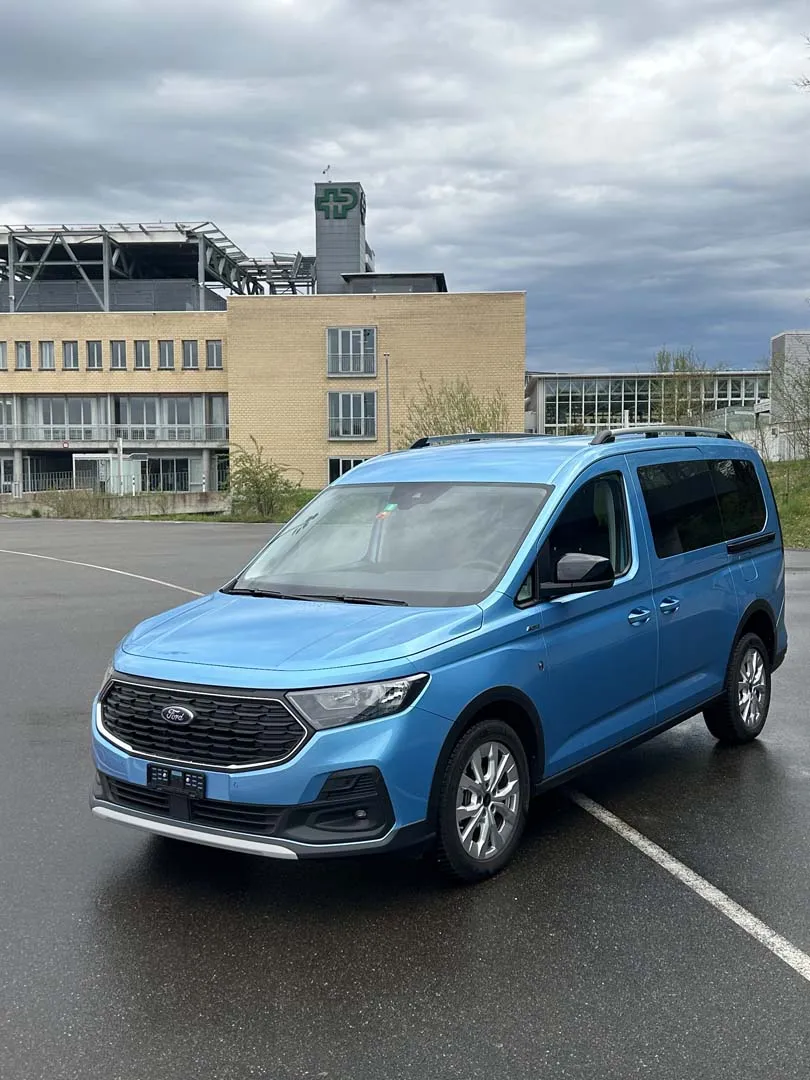 Blauer Ford Transit Connect-Minivan auf einem Parkplatz vor einem Gebäude unter bewölktem Himmel.