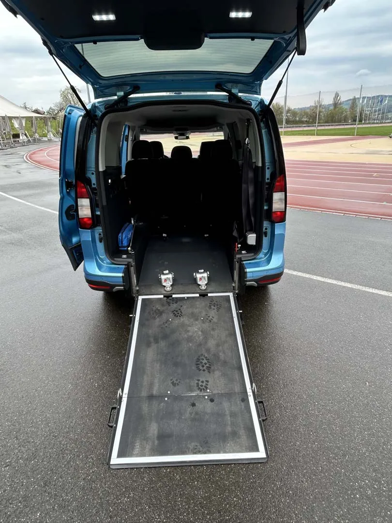 Blauer Minivan mit ausgefahrener Rollstuhlrampe am Heck auf einem Parkplatz.