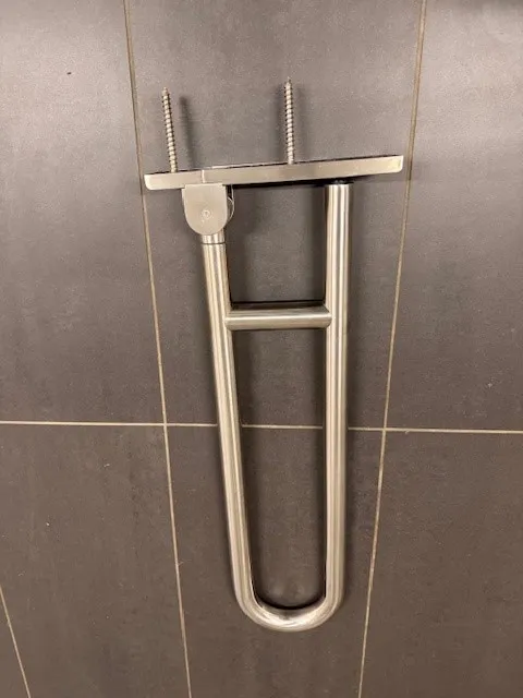 occasion-hewi-stuetzgriff-lippuner-03.jpg Klappbarer Stützgriff aus Metall an einer grauen Fliesenwand im Badezimmer montiert.