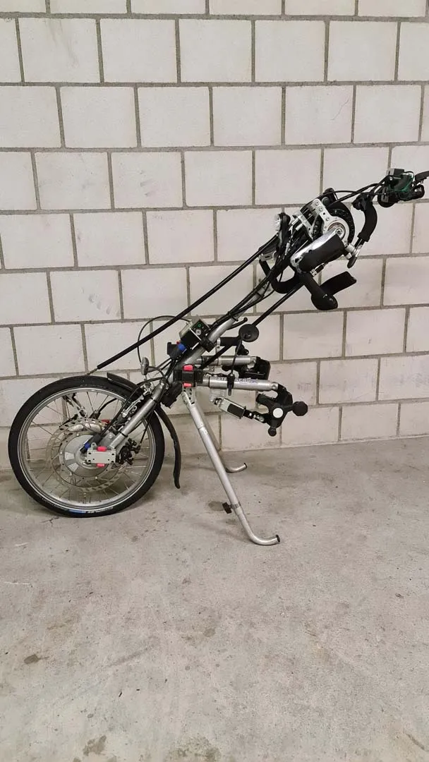 occasion-stricker-vorspannbike-nguyen-01.jpg Rollstuhlvorsatz mit Handkurbel an weißer Wand lehnend.