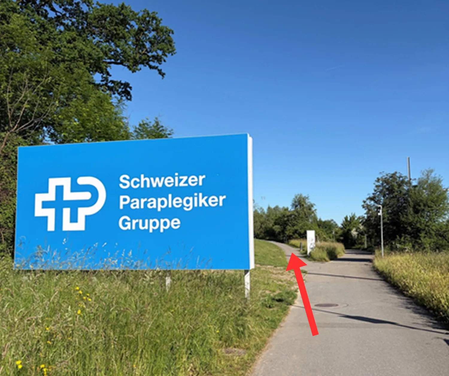 Blaues Schild der Schweizer Paraplegiker-Gruppe an einem Fußweg mit Bäumen im Hintergrund.