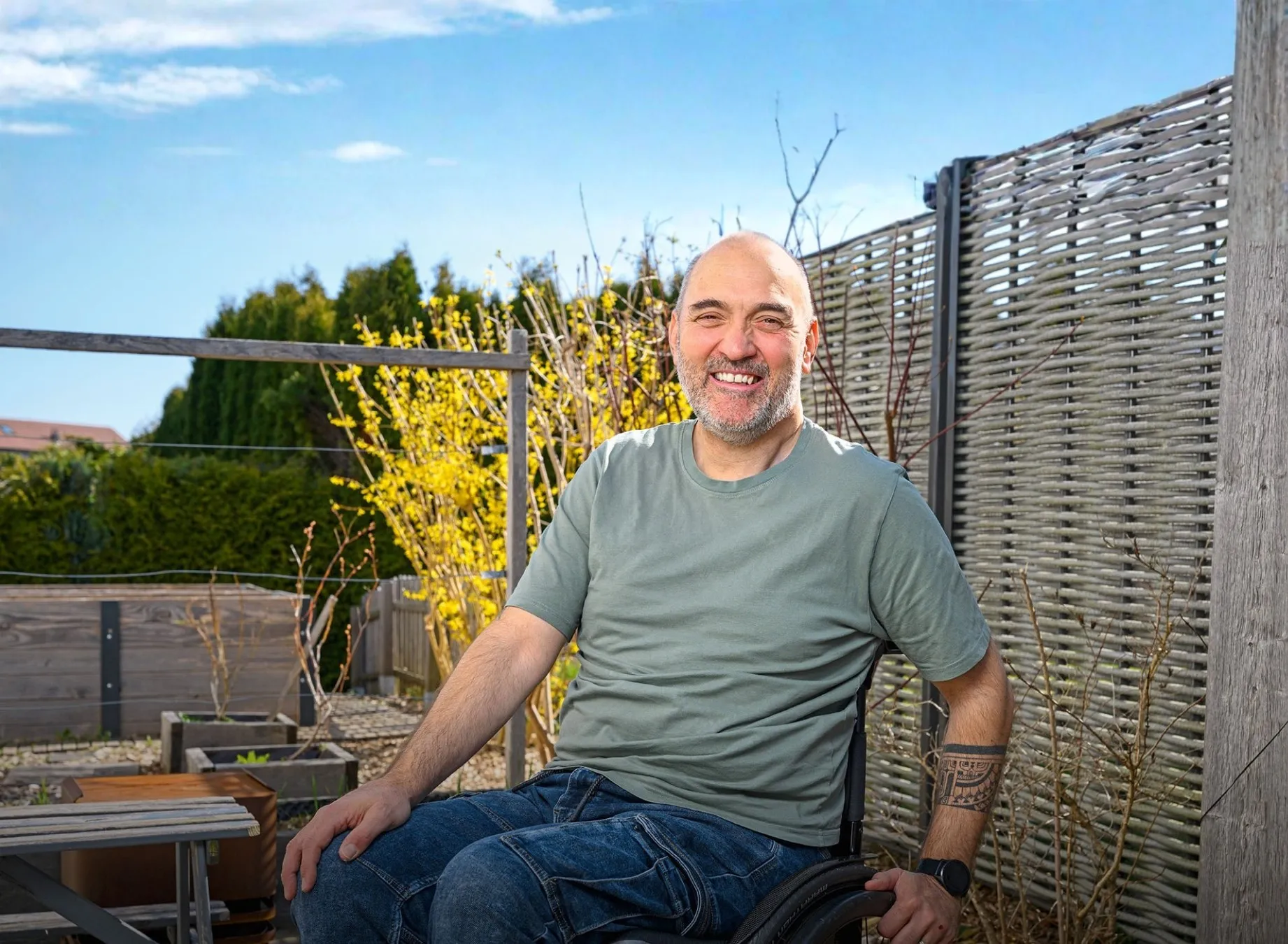 Lächelnder Mann im Rollstuhl sitzt bei sonnigem Wetter auf einer Terrasse im Garten – selbstbestimmtes Leben, Barrierefreiheit und Lebensfreude im Alltag.