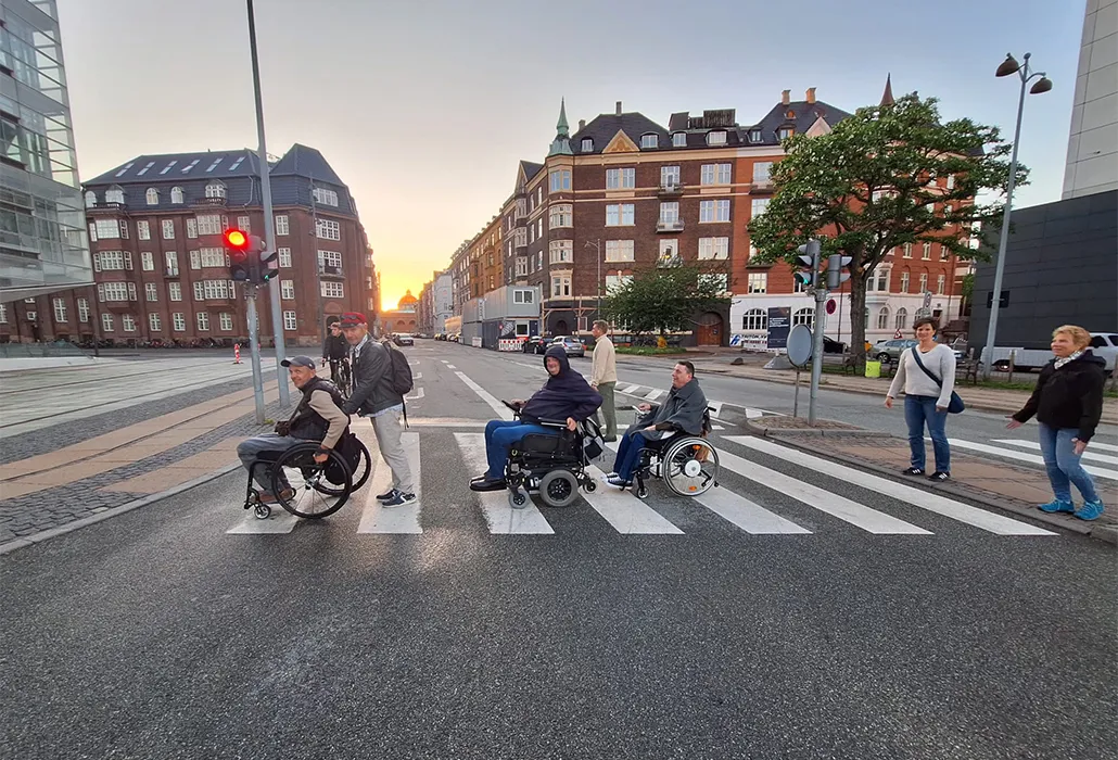 Das Bild zeigt barrierefreie Mobilität im öffentlichen Raum: Menschen mit und ohne Behinderung überqueren gemeinsam einen Fußgängerüberweg in einer modernen Stadt. Die Szene veranschaulicht inklusive Stadtentwicklung, zugängliche Infrastruktur, soziale Teilhabe und die Förderung von Selbstbestimmung für Rollstuhlnutzer im Alltag.