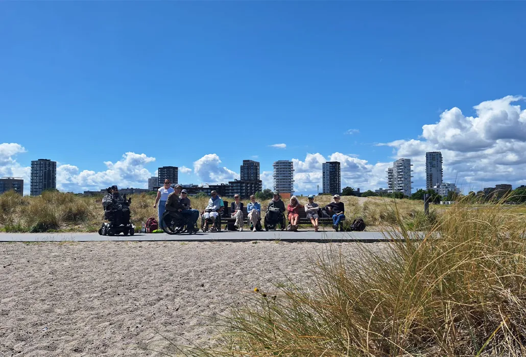 Menschen mit und ohne Mobilitätseinschränkungen verbringen gemeinsam Zeit auf einer barrierefrei zugänglichen Promenade in einer naturnahen Küstenlandschaft. Das Bild steht für Inklusion, Teilhabe, barrierefreien Tourismus und zugängliche Freizeitangebote im öffentlichen Raum.