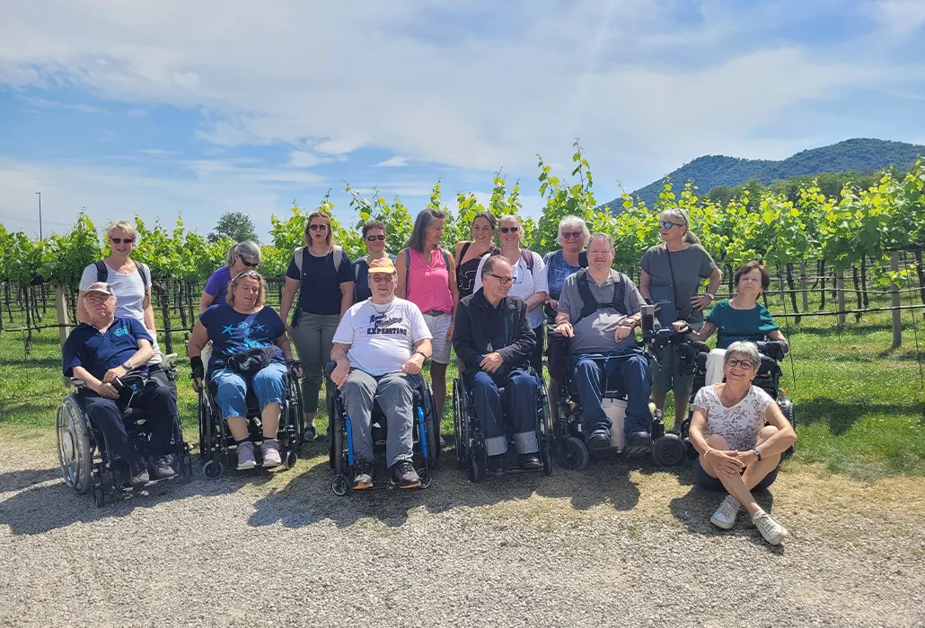 Eine Gruppe von Menschen mit und ohne Rollstuhl posiert bei sonnigem Wetter in einem Rebberg vor grünen Weinreben und Hügeln – barrierefreier Ausflug, Inklusion und Gemeinschaft in der Natur.