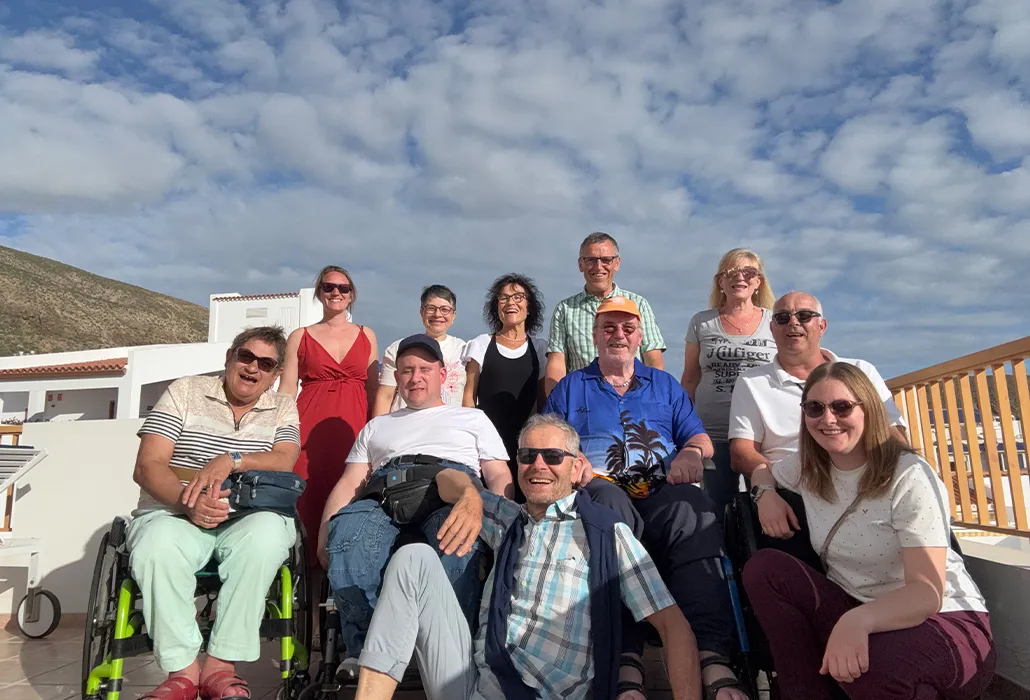 Eine lachende Reisegruppe mit Menschen mit und ohne Rollstuhl posiert auf einer sonnigen Terrasse – barrierefreie Ferien, Inklusion und gemeinsames Erlebnis in einer südlichen Ferienregion.