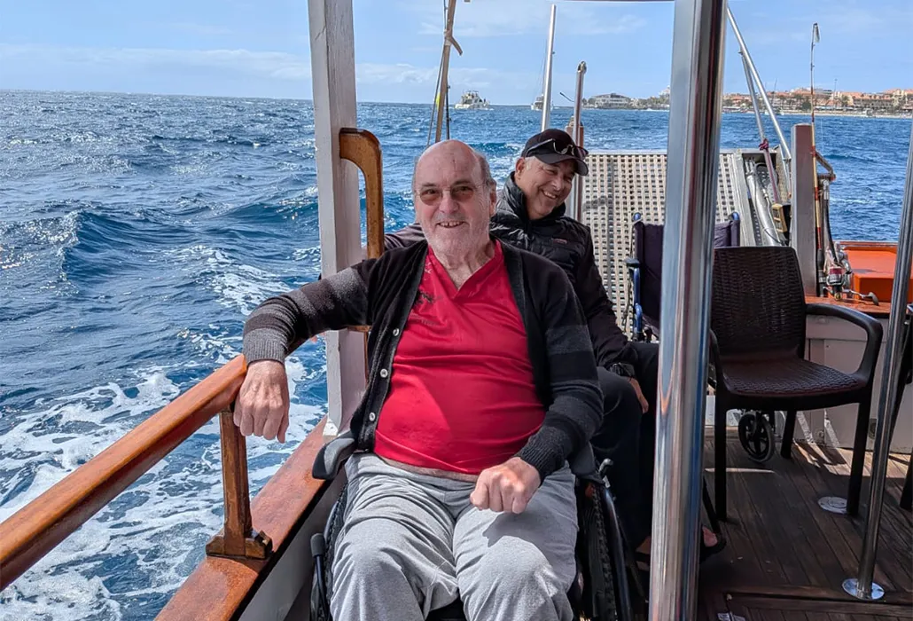 Zwei Männer im Rollstuhl geniessen eine sonnige Bootsfahrt auf dem Meer mit Blick auf die Küste – barrierefreie Reise, Inklusion und Ferienerlebnis am Wasser.